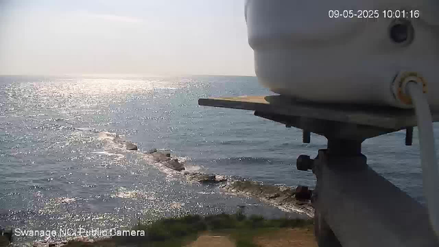 A view of a large body of water, possibly the ocean, with sunlight reflecting off its surface. In the foreground, part of a webcam device is visible, mounted on a pole or structure. The date and time displayed in the top right corner are 09-05-2025, 10:01:16. The scene appears sunny and calm with gentle waves.