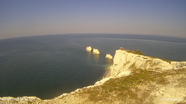 A view from a webcam showing white chalk cliffs extending into the sea with a small building and antenna on top of the cliffs. In the ocean, there are several large white rock formations or stacks near the cliffs. The sky is clear with a gradient from light blue to darker shades near the horizon. The water appears calm, and the scene is bathed in sunlight.