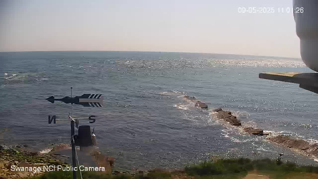 A view of the ocean with sunlight reflecting off the water, a rocky shoreline extending into the sea, and a clear sky above. There is a weather vane and a compass rose at the bottom left corner of the image, with the label "Swanage NCI Public Camera." The timestamp in the top right corner reads "00-05-2025 11:10:26."