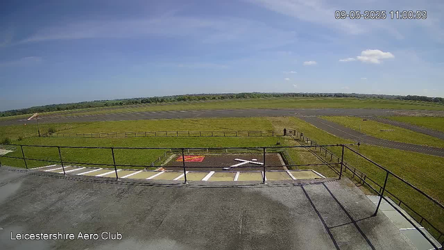 The image shows a view from a webcam at Leicestershire Aero Club. In the foreground, there is a paved area with a black railing, some empty parking spaces, and a small helipad marked with a white helicopter symbol and red rectangular markings. Beyond this, there is a grassy field enclosed by a wooden fence, leading to an open tarmac runway stretching across the middle of the image. In the background, there are gently rolling green fields with a distant tree line under a mostly clear sky with a few scattered clouds. The timestamp in the top right corner reads "09-05-2025 11:00:53."