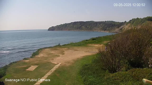 A view of a peaceful coastal landscape with a path leading to the beach. The shoreline is bordered by a grassy area and some bushes or small trees. In the distance, there is a large, tree-covered hill or cliff extending into the water. The sky is clear and calm, and a single person can be seen walking near the bushes on the right side. The date and time are displayed in the top right corner: September 5, 2025, at 12:00:52.