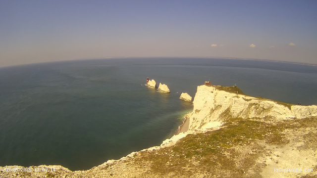 A view from a webcam shows white chalk cliffs extending into the sea, with a small lighthouse and a building on top of the cliffs. The ocean stretches out to the horizon under a clear sky with a few scattered clouds, and the sunlight illuminates the cliffs and the water.