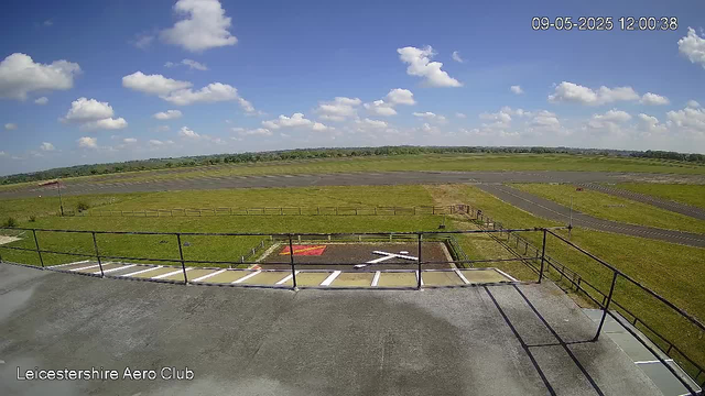 A view from a webcam showing an open airfield with a clear blue sky dotted with fluffy white clouds. There are grassy areas and paved runways extending into the distance. In the foreground, there is a helipad marked with a white "H" and a small, fenced parking space. The time and date are displayed in the top right corner as "09-05-2025 12:00:38." The bottom left corner shows the text "Leicestershire Aero Club."