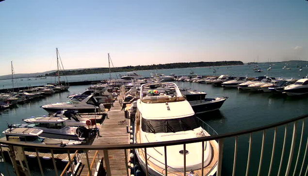 A marina filled with numerous boats docked along wooden piers, with sailboats and motorboats moored side by side. Calm water reflects the boats, and a distant landmass is visible under a clear sky. The photo is taken from an elevated perspective behind a curved metal railing.