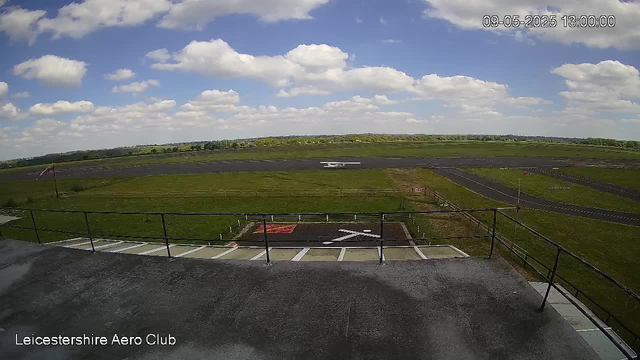 A view from a webcam showing a runway and surrounding grassy fields under a partly cloudy sky. In the distance, there is a small aircraft on the runway, and a windsock is visible on the left side. The timestamp in the upper right corner reads "09-05-2025 13:00:00," and at the bottom left, the text reads "Leicestershire Aero Club."