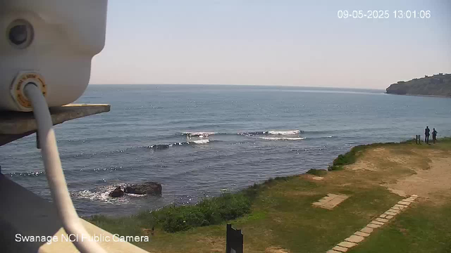 A scenic view of the ocean with gentle waves, a grassy area with a stone pathway leading toward the water, and a small group of three people standing near the shoreline on the right side. In the background, there is a distant landmass or cliff on the right. The weather appears clear and sunny. The timestamp in the top right corner reads "09-05-2025 13:01:06".