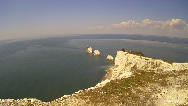 A wide view of the white chalk cliffs along a coastline, with one prominent lighthouse on a rock formation in the sea. The sky is mostly clear with some scattered clouds, and a small sailboat can be seen in the distance on the water.    
