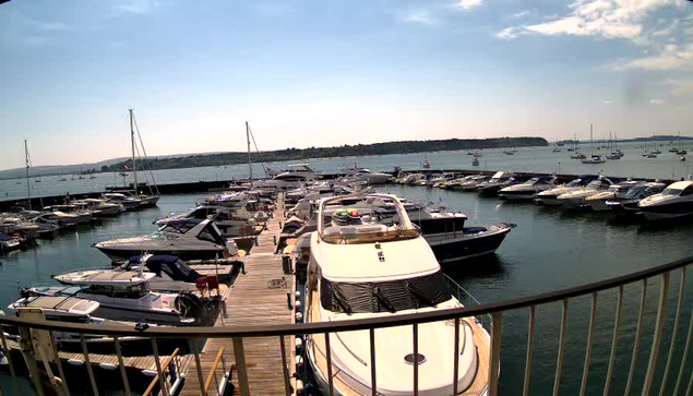 A marina filled with numerous boats and yachts docked along wooden piers. The water is calm, and the sky is partly cloudy with sunlight illuminating the scene. In the background, there is a low landmass or island extending across the horizon. A metal railing is visible in the foreground.