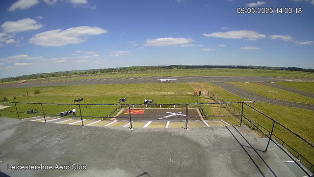 A view from a webcam at Leicestershire Aero Club on a bright, sunny day with scattered clouds. The foreground shows a concrete viewing platform with a metal railing. Below, there are small lawn areas with some parked motorcycles and people standing near them. In the distance, a small single-engine airplane is taxiing on the runway, and beyond that are open fields and the runway extending into the distance. The image includes a timestamp in the top right corner showing "09-05-2025 14:00:18".