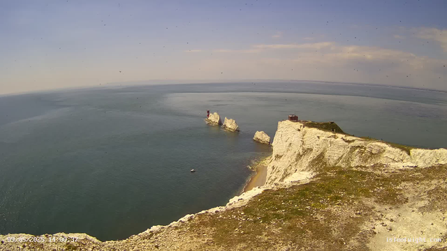 Bird's-eye view of white chalk cliffs and rock formations along a coastline, with the sea stretching out to the horizon. A small boat is visible near the shore, and a lighthouse stands on one of the rocks in the water. The sky is partly cloudy, and the overall scene is calm and scenic.