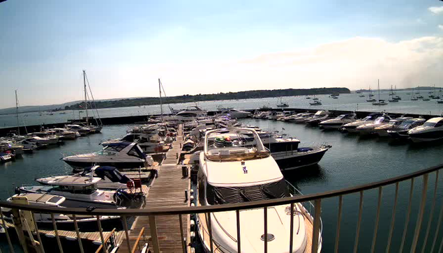 A marina filled with numerous boats and yachts docked along wooden piers on calm water, with a distant shoreline and a partly cloudy sky in the background.