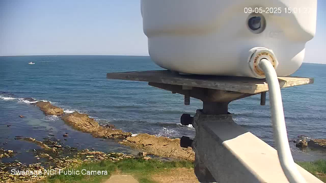 A webcam captures a view of the coastline with the ocean in the background. In the foreground, there is a close-up of a white device mounted on a metal and wooden stand, with a white cable connected to it. The coastline features rocky formations and some patches of green grass. The sky is clear, and a small boat is visible on the water in the distance. The timestamp shows the date as September 5, 2025, and the time as 15:01:22.