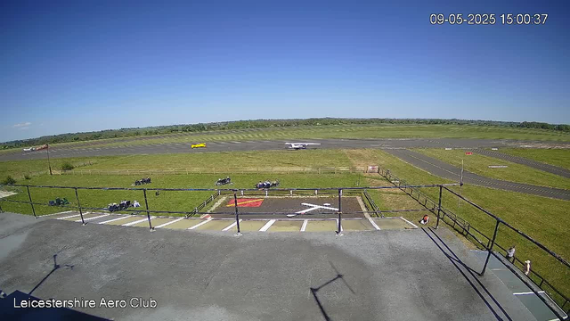 A view from a webcam at Leicestershire Aero Club showing the runway with a small aircraft taxiing in the background, a grassy area with several parked golf carts and a few people nearby, a fence surrounding the area, and a clear blue sky overhead. The timestamp indicates the date and time as September 5th, 2025, at 15:00:37.