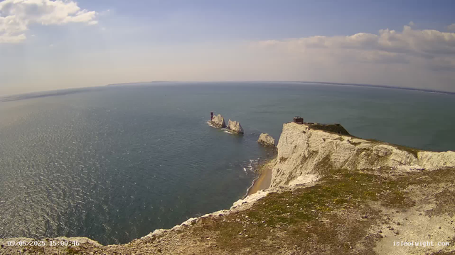 An aerial view of a coastal landscape showing white chalk cliffs extending into the sea. There are a few large rock formations just offshore and a lighthouse on one of the prominent cliffs. The water is a calm blue-green, and the sky above is partly cloudy with some white clouds scattered across a blue sky. The scene is captured in the late afternoon as indicated by the timestamp.