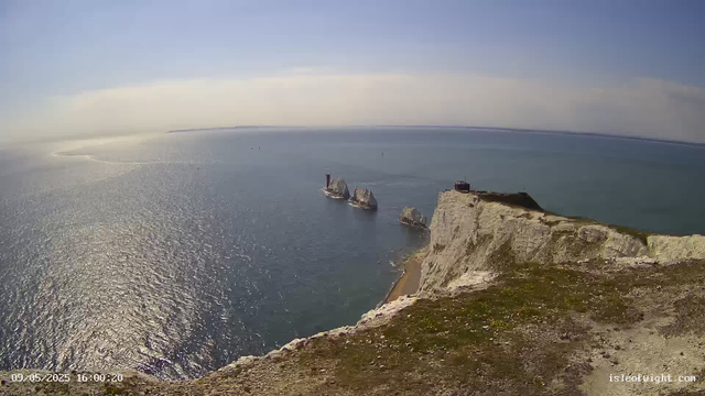 A panoramic view from a high cliff overlooking the ocean with white chalk cliffs in the foreground and several small rocky islands, including a lighthouse, in the distance. The sky is mostly clear with a few clouds, and the sunlight reflects off the water's surface. A timestamp in the bottom left corner reads "09/05/2025 16:00:20," and a website watermark "isleofight.com" is in the bottom right corner.