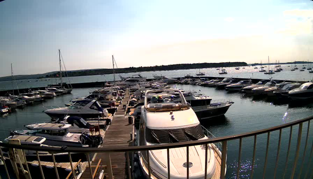A dock filled with various boats and yachts extends into a calm harbor, with more boats scattered across the water in the background. The sky is clear, and the horizon features a low, dark landmass. A curved railing is in the foreground.