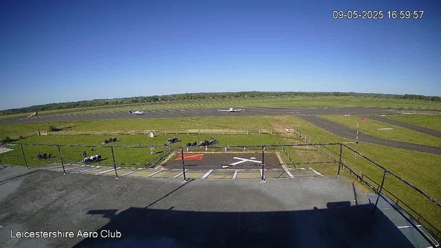 A clear view of an airfield at Leicestershie Aero Club on a sunny day. In the distance, two small aircraft are taxiing on the runway while another plane flies overhead. In the foreground, there is a small parking area with a few motorcycles and equipment, enclosed by a fence. The background features flat grassy fields under a bright blue sky. The date and time stamp in the top right corner reads "09-05-2025 16:59:57."