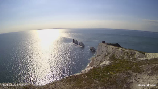A view of white chalk cliffs overlooking the ocean at sunset. The sun's reflection shimmers on the water, with three sea stacks rising from the sea near the cliffs. The sky is mostly clear with a few clouds near the horizon. The ground in the foreground is rocky and covered with patches of grass.