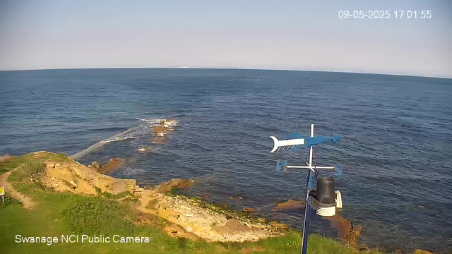 A view of a rocky coastline with green grass in the foreground, and the ocean extending to the horizon. A weather station with a wind vane and anemometer is visible near the shore. The sky is clear, and the image is timestamped "09-05-2025 17:01:55" in the top right corner.