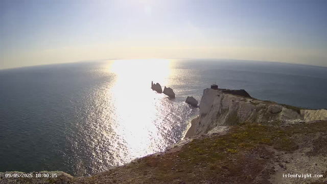 A view of the white chalk cliffs and the sea at sunset, with rocks and a small stone building on top of the cliff. The date and time displayed are 09/05/2025 18:00:05, and the website is isleofwight.com.