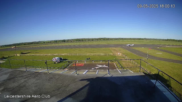 An outdoor scene of a small airport runway during daytime, with a parked vehicle and a dog on a grassy area in the foreground. A small aircraft is taxiing on the runway in the distance. The sky is clear and blue. The timestamp in the upper right corner reads "09-05-2025 18:00:13," and the location tag at the bottom left indicates "Leicestershire Aero Club."