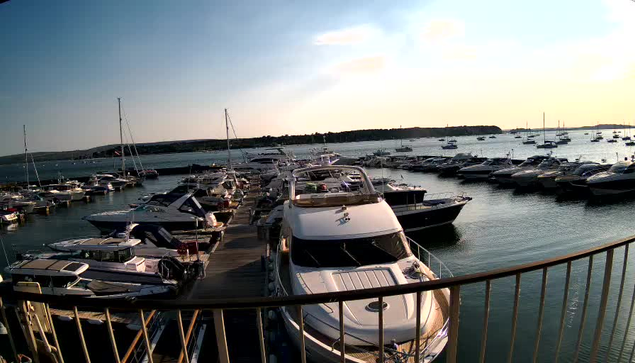 A view of a marina with multiple boats and yachts docked along wooden piers. The water is calm, and more boats are visible in the distance, extending out into a larger body of water. The sky is partly cloudy with the sun setting or rising, casting a warm glow on the scene. A curved railing is visible in the foreground.