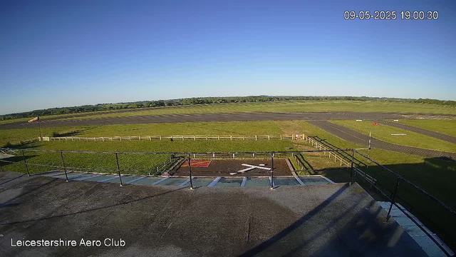 A view from a webcam at Leicestershire Aero Club showing a clear blue sky above a grassy and paved area of the airfield. The time and date are displayed in the top right corner as 09-05-2025 19:00:30. The foreground includes a part of a rooftop or platform with safety railings. The landscape stretches out into flat open fields beyond the airfield.