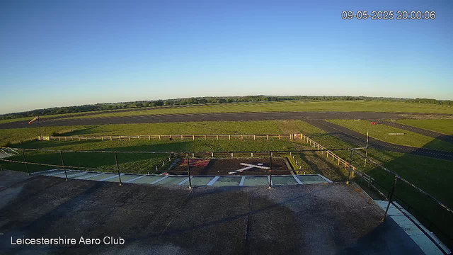 An image from a webcam showing an airfield with a wide open grassy area, taxiways, and runway extending into the distance under a clear blue sky. A small flag is visible on the left side near a fenced area. The scene is brightly lit, suggesting it is either early morning or late afternoon. The timestamp in the upper right corner reads "09-05-2025 20:00:06." The bottom left corner displays the text "Leicestershire Aero Club."