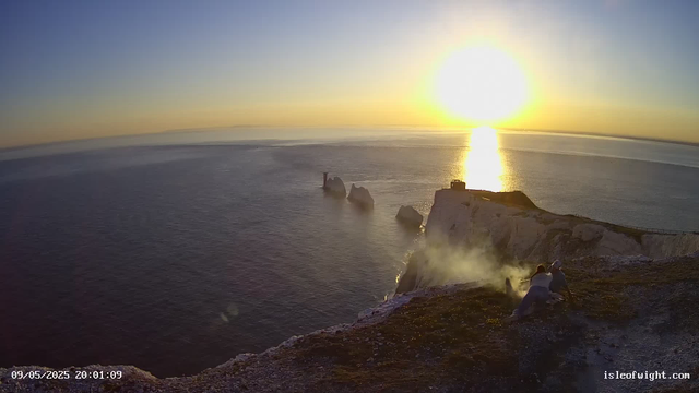 A view of the ocean at sunset with rocky cliffs and steam rising from the ground, with two people sitting near the edge of the cliff. The sun is low on the horizon, reflecting brightly on the water.