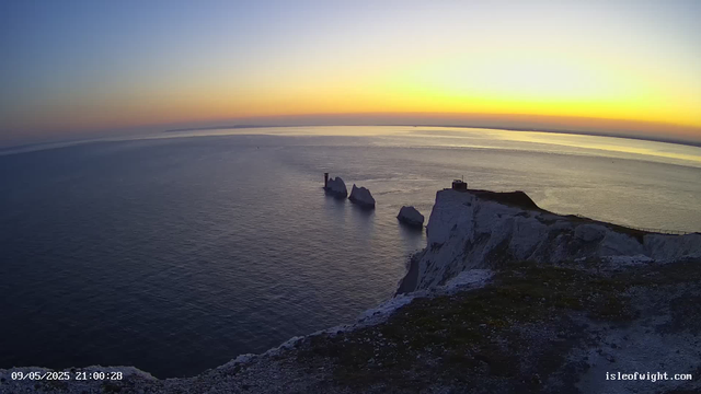 A scenic view of white chalk cliffs overlooking the ocean at sunset. The sky displays a gradient of warm colors from yellow to dark blue, with the sun setting on the horizon. There are several large rock formations in the water near the base of the cliffs, and a lighthouse along with a small building situated on the cliff top. The time stamp in the bottom left corner reads "09/05/2025 21:00:28," and the bottom right corner indicates the location as "isleofwight.com."