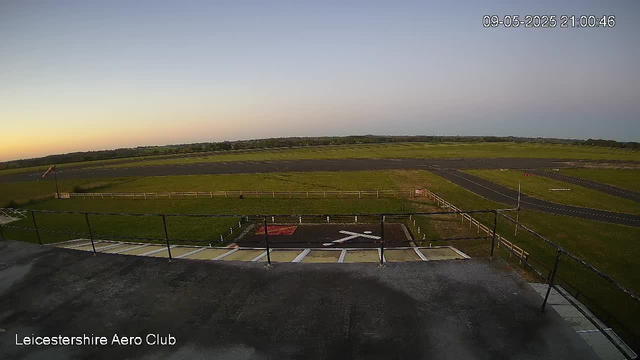 A view of an airfield at dusk, showing a small helicopter landing pad marked with a white helicopter symbol and a red and white border. The runway stretches into the distance with a few small runways or taxiways visible. There's a green grassy area surrounded by a white fence, and a black railing in the foreground. The sky transitions from light blue to darker shades as the sun has set. The timestamp in the top right corner reads "09-05-2025 21:00:46". The bottom left corner has the text "Leicestershire Aero Club."