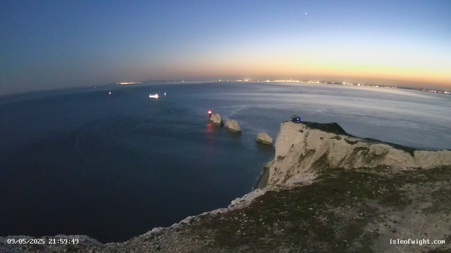 A view from a webcam showing a coastal scene at dusk or dawn. White cliffs extend into the water, with a small blue structure on top of one of the cliffs. The sea appears calm, with several boats visible in the water, some with lights on. The sky transitions from light near the horizon to darker blue overhead, with the faint glow of sunset or sunrise on the horizon. A bright star or planet is visible in the sky above the horizon. The timestamp on the image reads 09/05/2025 21:59:49, and the website isleofwight.com is displayed in the bottom right corner.