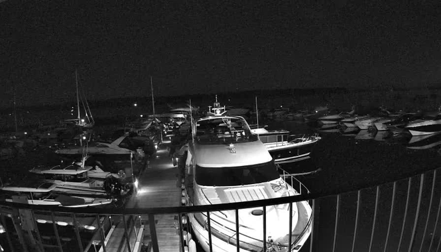 A black-and-white image of a harbor filled with various boats and yachts docked at night, viewed from a balcony or pier with a railing in the foreground. The scene is dark, with some boat lights faintly visible in the distance.