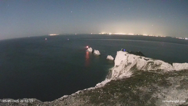 A nighttime view of white chalk cliffs and rock formations extending into the dark sea. There are a few lights on the cliffs and rocks, with some reflections on the water. The horizon shows a distant coastline with numerous city lights under a clear sky with a few visible stars. The timestamp in the bottom left corner reads "09/05/2025 22:59:59" and the website "isleofwight.com" is in the bottom right corner.