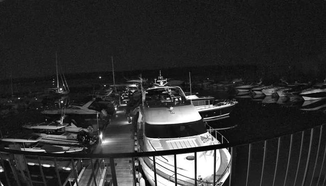 A black-and-white image from a webcam shows a marina at night with numerous boats docked. The boats are lined up along the piers, and the scene is dark, with only the boats and some reflections faintly visible. The railing of a balcony or deck is visible in the foreground.