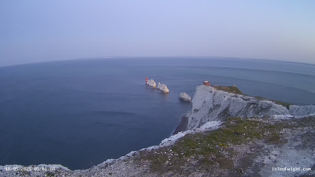 A view of white chalk cliffs along the coastline with large rock formations in the sea nearby. There is a lighthouse with a red top and a small building on the cliff edge. The ocean is calm and stretches out into the horizon under a clear sky. The timestamp reads "10/05/2025 05:01:08" and the website "isleofwight.com" is displayed at the bottom right corner.
