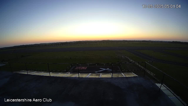 A view from a webcam overlooking an airfield during sunset or sunrise with a clear sky. The horizon is visible in the distance, and the foreground shows part of a rooftop or platform with safety railings. There is a helipad with an "H" marking and surrounding grassy areas. The timestamp in the top right corner indicates the date and time as 10-05-2025 at 05:01:04. The bottom left corner has the text "Leicestershire Aero Club."