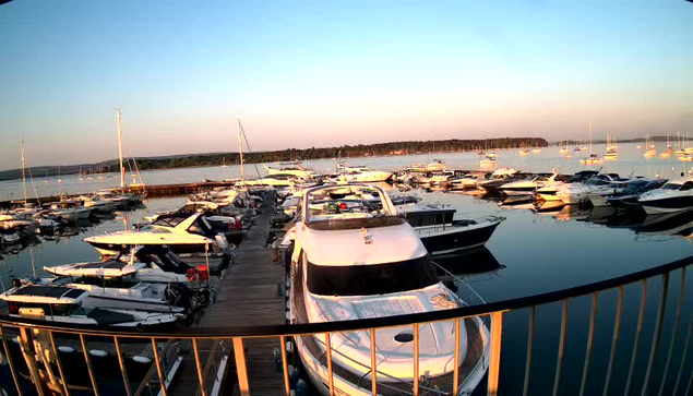 A marina filled with various boats and yachts anchored at wooden docks during sunset, with a calm body of water reflecting the vessels and a distant tree-lined shoreline under a clear sky.