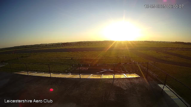 A wide view of an open field at sunrise, with the sun low on the horizon and sunlight casting long shadows. The foreground features a platform or terrace with metal railings, and the background shows vast, flat grassland extending to the horizon. The date and time are displayed in the upper right corner. The bottom left corner has the text "Leicestershire Aero Club."