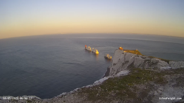 A view of white chalk cliffs rising sharply from the ocean, with three prominent sea stacks nearby. The ocean is calm, and the sky is clear with a soft, warm light suggesting either sunrise or sunset. The top of the cliffs is green with a small structure visible on the right. The timestamp in the bottom left corner indicates the date and time as October 5, 2025, at 6:00 AM.
