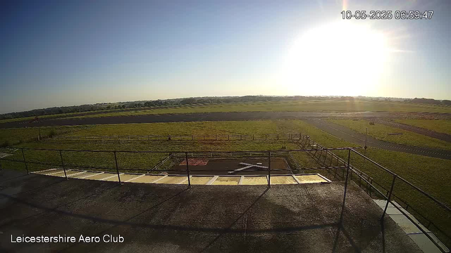 A wide view of a grassy airfield area during daylight, with a clear sky and bright sunlight in the distance. The foreground features a rooftop or platform with metal railings, and a fenced perimeter surrounds a section of the grass. A paved road runs across the right side of the image. There are no people or planes visible. The timestamp in the top right corner shows "10-05-2025 06:59:47," and the text "Leicestershire Aero Club" is displayed at the bottom left corner.