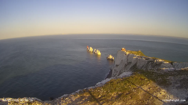 A scenic view of white chalk cliffs overlooking the ocean at sunset. In the distance, there is a prominent stack with a lighthouse on top, and a small building further along the cliff edge. The sky is clear with a gentle gradient from light near the horizon to darker blue higher up. The foreground shows the rocky, grassy edge of the cliff.