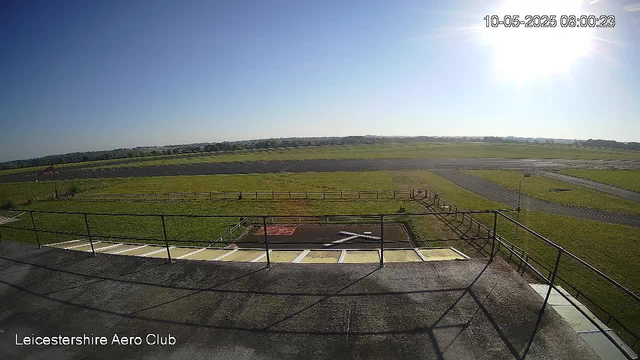 A view from a webcam showing an open grassy area with a small paved section and a fence. In the background, there are runways and open fields under a bright, clear sky with the sun shining. The image is labeled "Leicestershire Aero Club" in the bottom left corner, and the timestamp in the top right corner reads "10-05-2025 08:00:23."