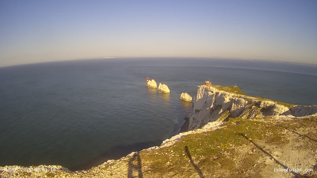 A view of white chalk cliffs extending into the sea with a lighthouse and a small building at the edge. Several large rock formations are visible offshore, and the sky is clear with a distant horizon.