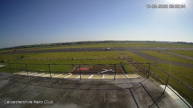 An aerial view of Leicestershire Aero Club showing a runway, grassy fields, and an aircraft in the distance under a clear blue sky with bright sunlight. The timestamp in the upper right corner reads 10-05-2025 09:00:34.