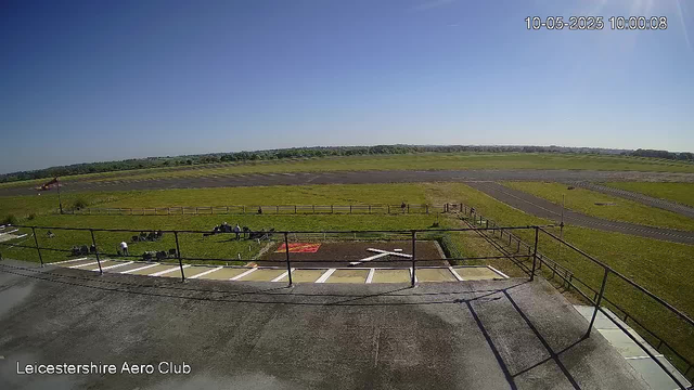 An outdoor view from a webcam at Leicesterhire Aero Club on a clear, sunny day. The foreground shows a parking area with white lines and a railing, and a few people sitting or standing on the grass nearby. Beyond the parking lot, there is a grass field with a small red mat, and further back, runways and open grassy spaces stretch into the distance under a blue sky. The timestamp in the top right corner reads "10-05-2025 10:00:08".