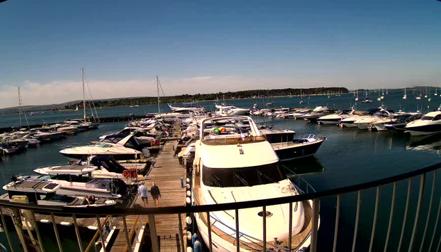 A marina filled with numerous boats and yachts docked along wooden piers. The boats vary in size, some large and some small, with a clear blue sky overhead and calm water surrounding the docks. In the background, there is a distant shoreline with low hills or landmasses. A few people are walking along the pier.