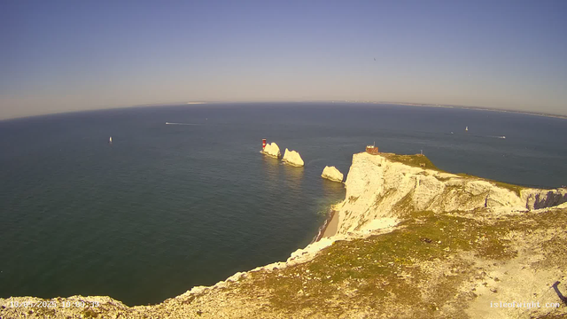 A panoramic view of white chalk cliffs along a coastline, with a small lighthouse on a grassy area at the edge. The cliffs jut out into the sea, with some large rocks near the base. The sea is calm with a few sailboats and boats visible in the distance under a clear blue sky.