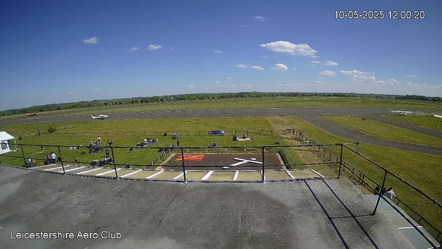 A clear day at Leicestershire Aero Club with a bright blue sky and a few scattered clouds. The foreground shows a terrace with black railings and a concrete floor, where a few people are sitting or standing. Below, a grassy area has multiple people sitting or lying on the grass, some next to small aircraft parked on the grass. Beyond that, a runway extends into the distance with a couple of small planes parked alongside. The landscape is flat and open, with a line of trees visible on the horizon. The timestamp in the top right corner reads "10-05-2025 12:00:20."
