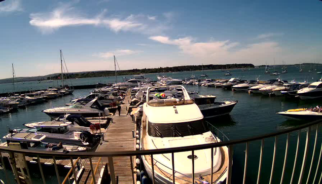 A marina with numerous boats and yachts docked along wooden piers. The water is calm and blue, with boats of various sizes moored on either side. The sky is partly cloudy with streaks of white clouds. In the background, there is a distant shoreline covered with greenery.